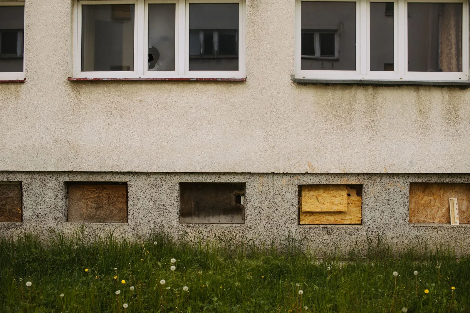 Exterior view of an abandoned building with boarded windows and overgrown grass.