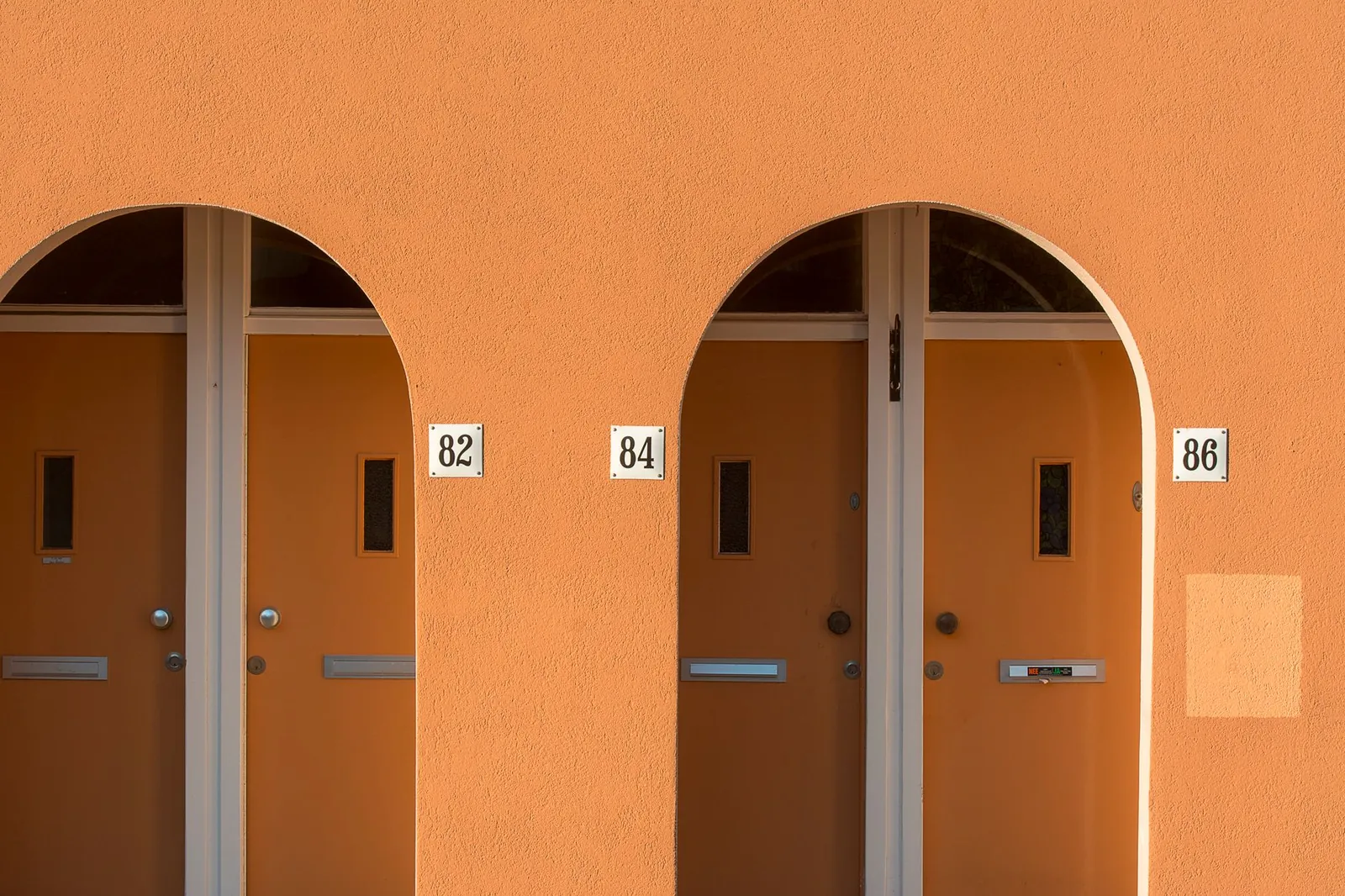 Stylish orange doors with arched frames and house numbers 82, 84, 86 on a sunny wall.