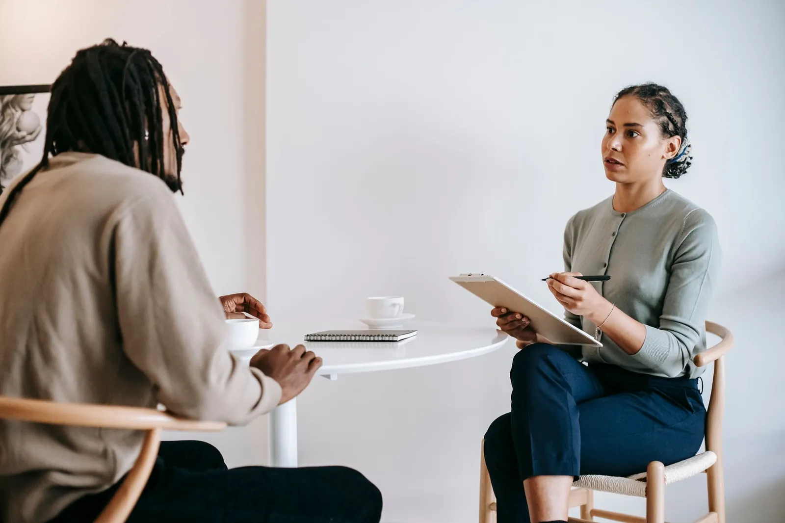Focused ethnic female interviewer or psychotherapist asking questions and writing notes in clipboard while looking at African American male